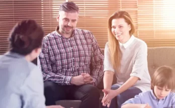 A family sits together in a cozy room with blinds, engaged in a happy discussion with another person about parenting.