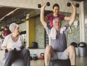 A senior man lifts weights while seated on a gym ball, assisted by a trainer, with another older woman working out beside them.