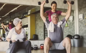 A senior man lifts weights while seated on a gym ball, assisted by a trainer, with another older woman working out beside them.