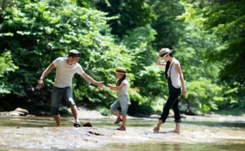A family enjoys a sunny day by a river, wading through the water, with the father helping the daughter while the mother walks alongside.