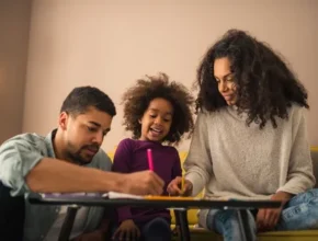 The parents sit on a couch, engaged in creative activities with their son at a table with colored markers and paper in a cozy, well-lit room.