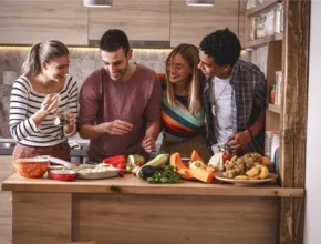 A group of four friends gathers around a wooden kitchen island, preparing fresh fruits and vegetables together, surrounded by bright ingredients.