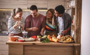 A group of four friends gathers around a wooden kitchen island, preparing fresh fruits and vegetables together, surrounded by bright ingredients.