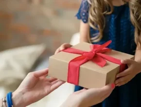 A child in a blue dress excitedly receives a gift box with a red ribbon from an adult's hands.