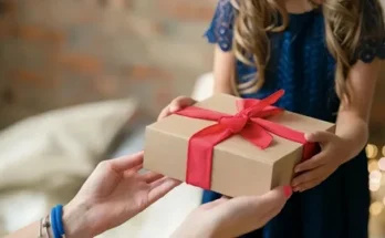 A child in a blue dress excitedly receives a gift box with a red ribbon from an adult's hands.