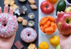 A person holds a sprinkle-covered donut in one hand and a red apple in the other, surrounded by various fruits and snacks.