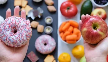 A person holds a sprinkle-covered donut in one hand and a red apple in the other, surrounded by various fruits and snacks.