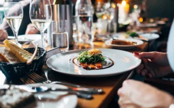 An elegantly set dining table with sparkling wine, water, and a gourmet plate of salmon topped with greens, alongside fresh bread.