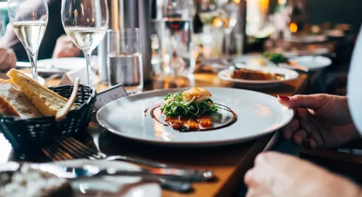 An elegantly set dining table with sparkling wine, water, and a gourmet plate of salmon topped with greens, alongside fresh bread.