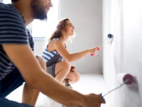 A woman paints a wall with a roller while a man sits nearby, both engaged in home improvement. Natural light fills the room.