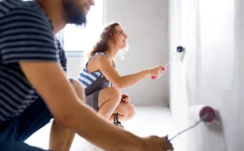 A woman paints a wall with a roller while a man sits nearby, both engaged in home improvement. Natural light fills the room.
