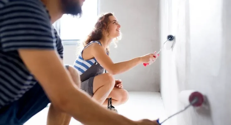 A woman paints a wall with a roller while a man sits nearby, both engaged in home improvement. Natural light fills the room.