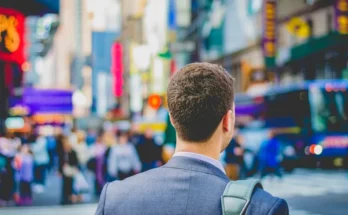 A suited man stands in Times Square, looking toward a bustling crowd and vibrant advertisements, capturing the city's energetic atmosphere.