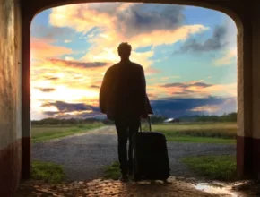 A silhouetted figure with a suitcase stands in an archway, gazing at a vibrant sunset over a rural landscape.