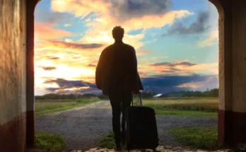 A silhouetted figure with a suitcase stands in an archway, gazing at a vibrant sunset over a rural landscape.