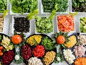 A vibrant array of fresh vegetables and toppings neatly arranged in bowls, featuring greens, peppers, olives, and fruits.