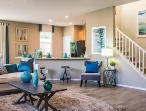 Bright, modern living room with beige walls, cozy furniture, and teal accents. Opens to a kitchen and staircase in the background.