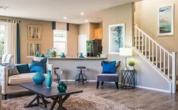 Bright, modern living room with beige walls, cozy furniture, and teal accents. Opens to a kitchen and staircase in the background.