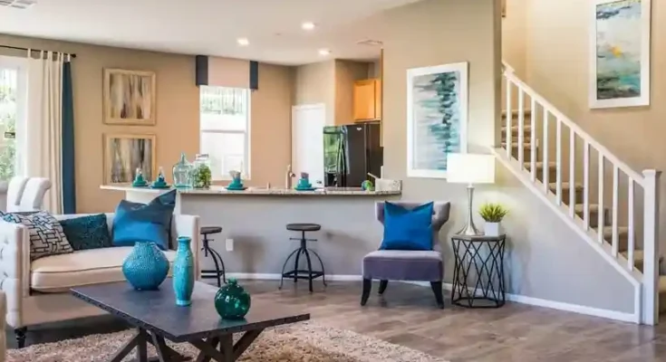 Bright, modern living room with beige walls, cozy furniture, and teal accents. Opens to a kitchen and staircase in the background.