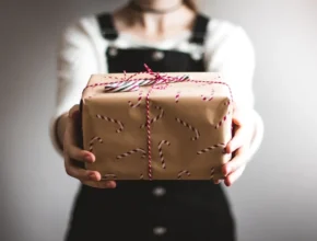 A person in a white shirt and black overalls holds out a gift wrapped in brown paper with a red and white string and candy cane pattern.