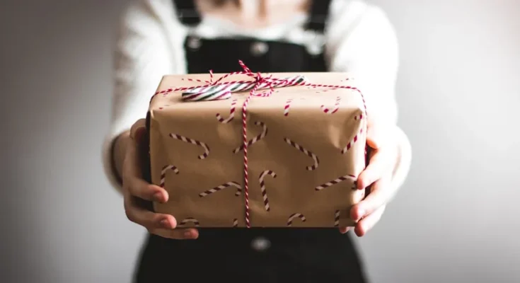 A person in a white shirt and black overalls holds out a gift wrapped in brown paper with a red and white string and candy cane pattern.