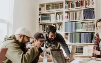 A group of people collaborate around a table in a library, surrounded by bookshelves filled with various books and folders.