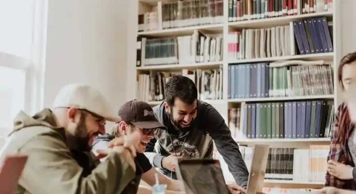 A group of people collaborate around a table in a library, surrounded by bookshelves filled with various books and folders.