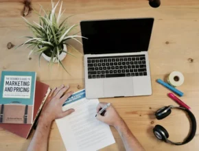 An overhead view of a workspace with a laptop, plant, books on marketing, colored markers, tape, headphones, and a person writing.