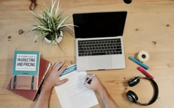 An overhead view of a workspace with a laptop, plant, books on marketing, colored markers, tape, headphones, and a person writing.