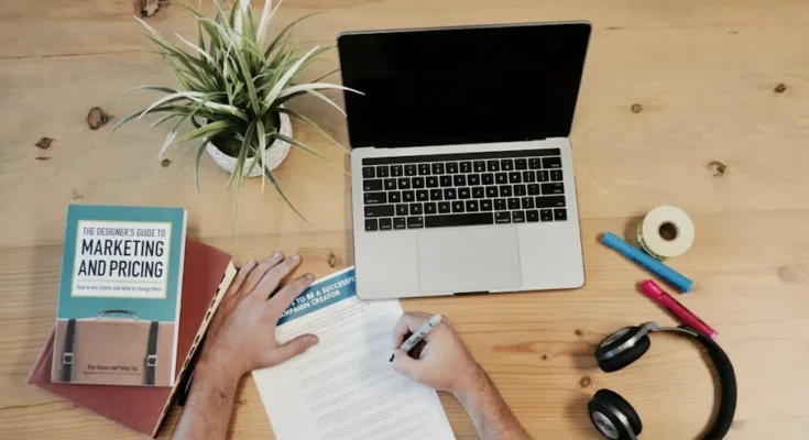 An overhead view of a workspace with a laptop, plant, books on marketing, colored markers, tape, headphones, and a person writing.