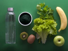 A collection of fresh fruits and vegetables: a bottle of water, spirulina powder, lettuce, banana, kiwi, egg, and green apple on a green background.