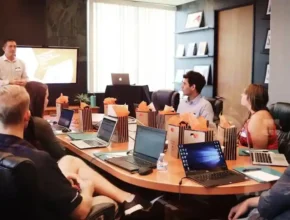 A meeting room with several attendees around a large table, laptops open, and gift bags prominently displayed, while a presenter speaks.
