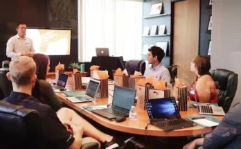 A meeting room with several attendees around a large table, laptops open, and gift bags prominently displayed, while a presenter speaks.