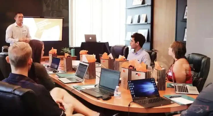 A meeting room with several attendees around a large table, laptops open, and gift bags prominently displayed, while a presenter speaks.