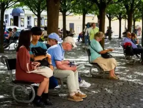 A lively public square with people sitting on benches under leafy trees, enjoying their surroundings and engaged in various activities.