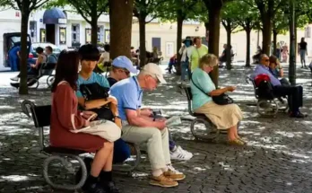 A lively public square with people sitting on benches under leafy trees, enjoying their surroundings and engaged in various activities.