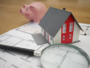 A model house on blueprints, accompanied by a magnifying glass and a pink piggy bank, symbolizing home planning and finance.