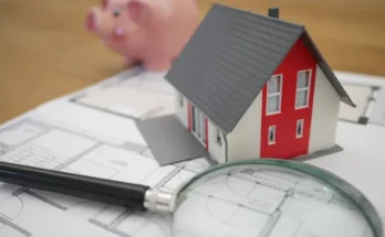 A model house on blueprints, accompanied by a magnifying glass and a pink piggy bank, symbolizing home planning and finance.