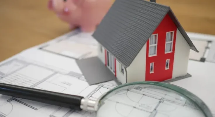 A model house on blueprints, accompanied by a magnifying glass and a pink piggy bank, symbolizing home planning and finance.