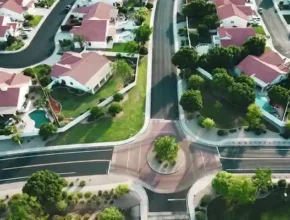 Aerial view of suburban neighborhood with houses, green lawns, and a circular intersection surrounded by trees and landscaped areas.