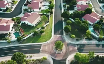 Aerial view of suburban neighborhood with houses, green lawns, and a circular intersection surrounded by trees and landscaped areas.