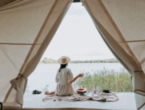 A woman in a straw hat sits by a lakeside, sipping a drink, with a picnic spread on a blanket seen through a tent opening.