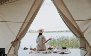 A woman in a straw hat sits by a lakeside, sipping a drink, with a picnic spread on a blanket seen through a tent opening.