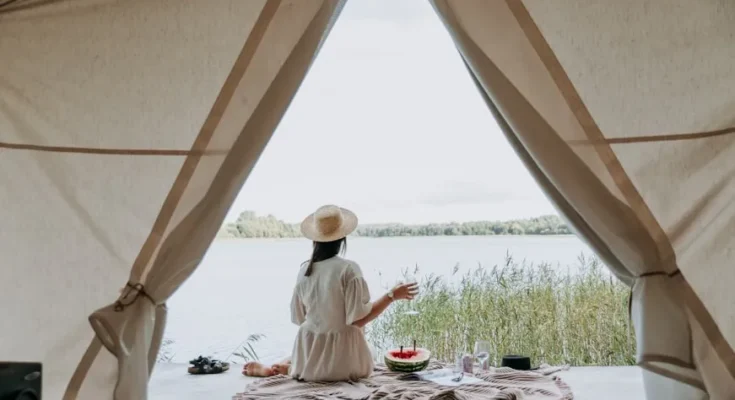 A woman in a straw hat sits by a lakeside, sipping a drink, with a picnic spread on a blanket seen through a tent opening.