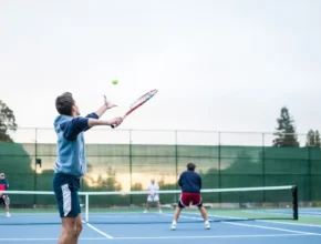 A player serves a tennis ball on a court, while two opponents prepare to receive. Trees and a cloudy sky are visible in the background.