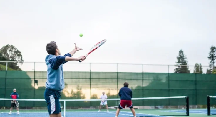 A player serves a tennis ball on a court, while two opponents prepare to receive. Trees and a cloudy sky are visible in the background.