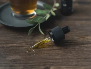A dropper bottle with golden oil lies on a wooden surface beside a cup of tea and green leaves, creating a calming, natural vibe.