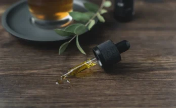 A dropper bottle with golden oil lies on a wooden surface beside a cup of tea and green leaves, creating a calming, natural vibe.