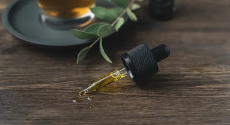 A dropper bottle with golden oil lies on a wooden surface beside a cup of tea and green leaves, creating a calming, natural vibe.