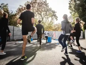 A group of individuals jogs together outdoors, with trees and sunlight in the background, engaged in a fitness workout.